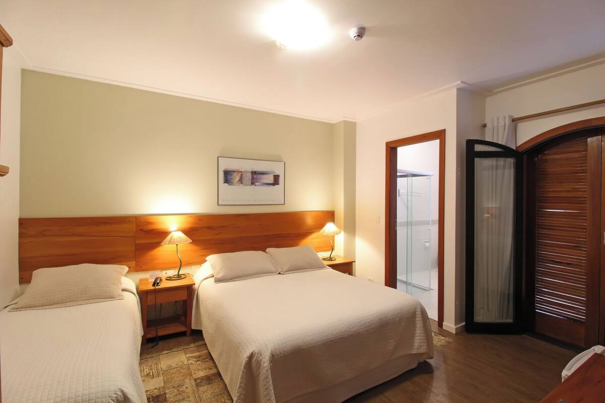 Une chambre d’hôtel avec deux lits, une tête de lit en bois, une table de chevet avec une lampe, un tableau encadré au mur et une porte coulissante avec un rideau.