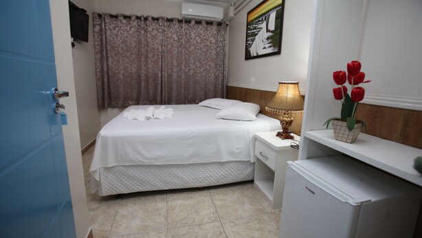Ein Hotelzimmer mit Bett, Nachttisch, Lampe, kleinem Kühlschrank und einem Topf mit roten Blüten.