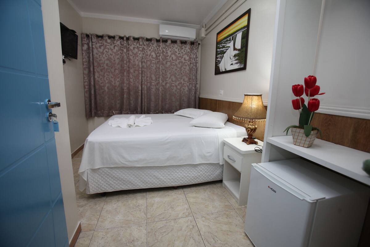 A hotel room with a bed, a nightstand, a lamp, a small refrigerator, and a potted plant with red flowers.