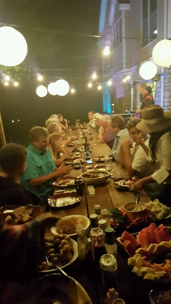 Ryhmä ihmisiä istuu pitkän ruokapöydän ympärillä, ja pöydällä on erilaisia ruokia ja juomia.