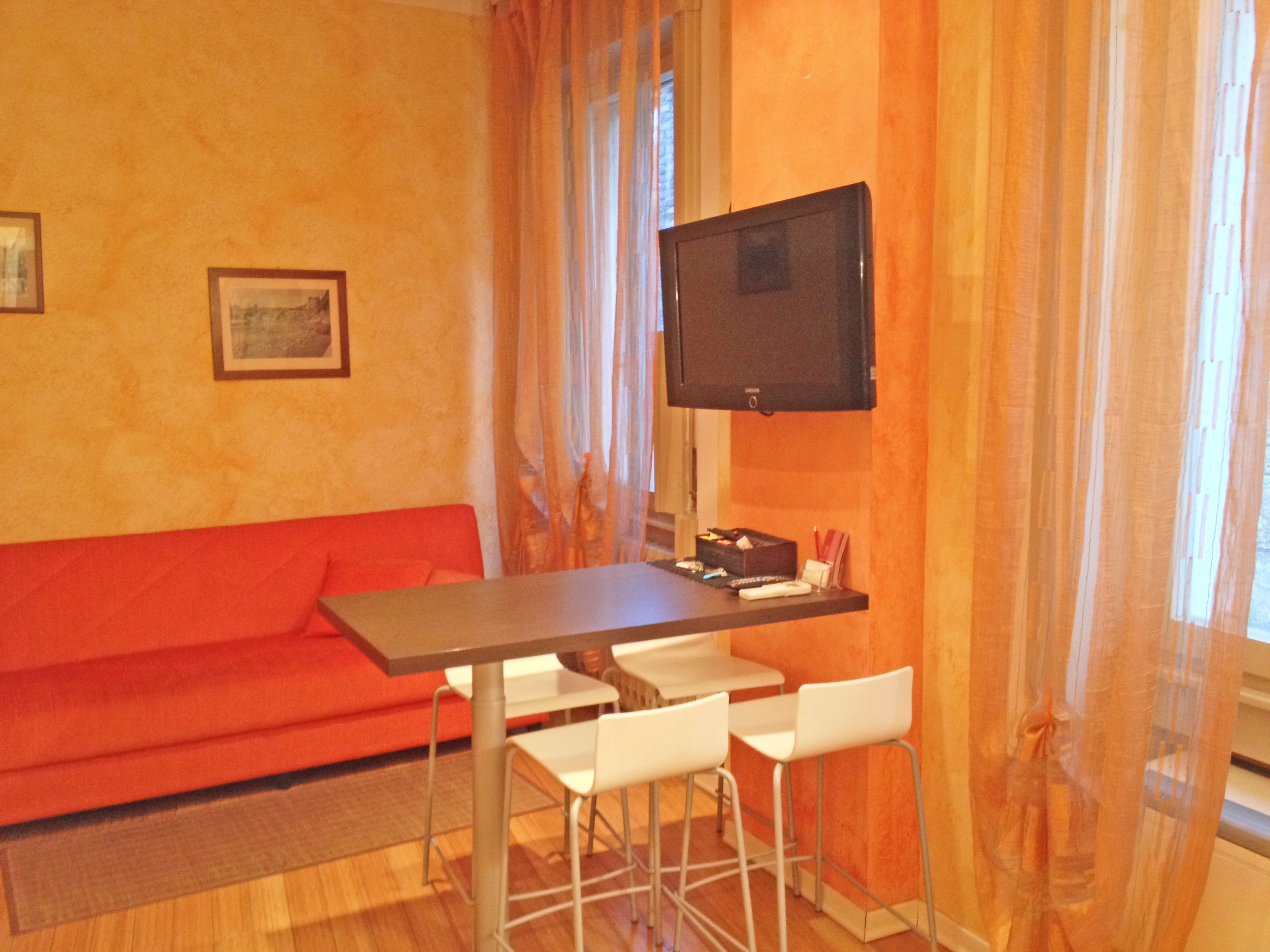 A room with a red sofa, a small table with two chairs, a television mounted on the wall, and a window with sheer curtains.