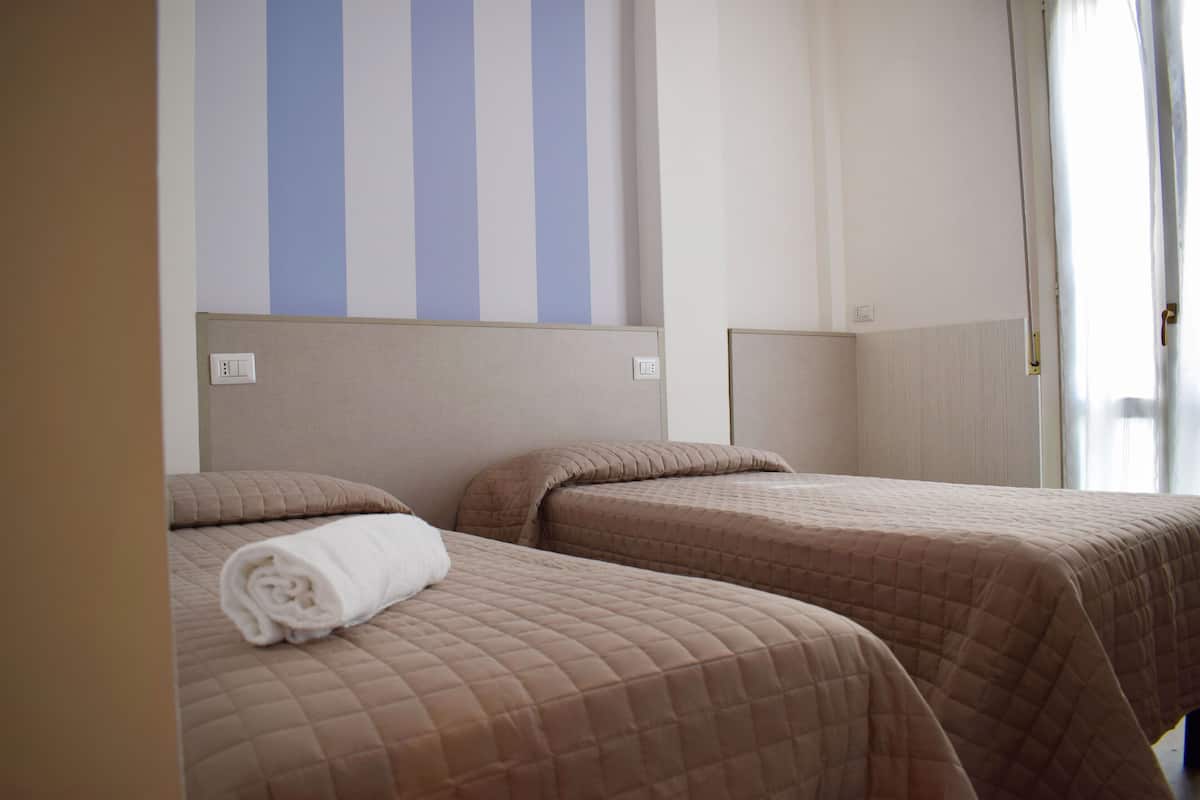 A hotel room with two single beds, each with a quilted brown bedspread and a rolled white towel. The room has a striped wall design and a door with a curtain.