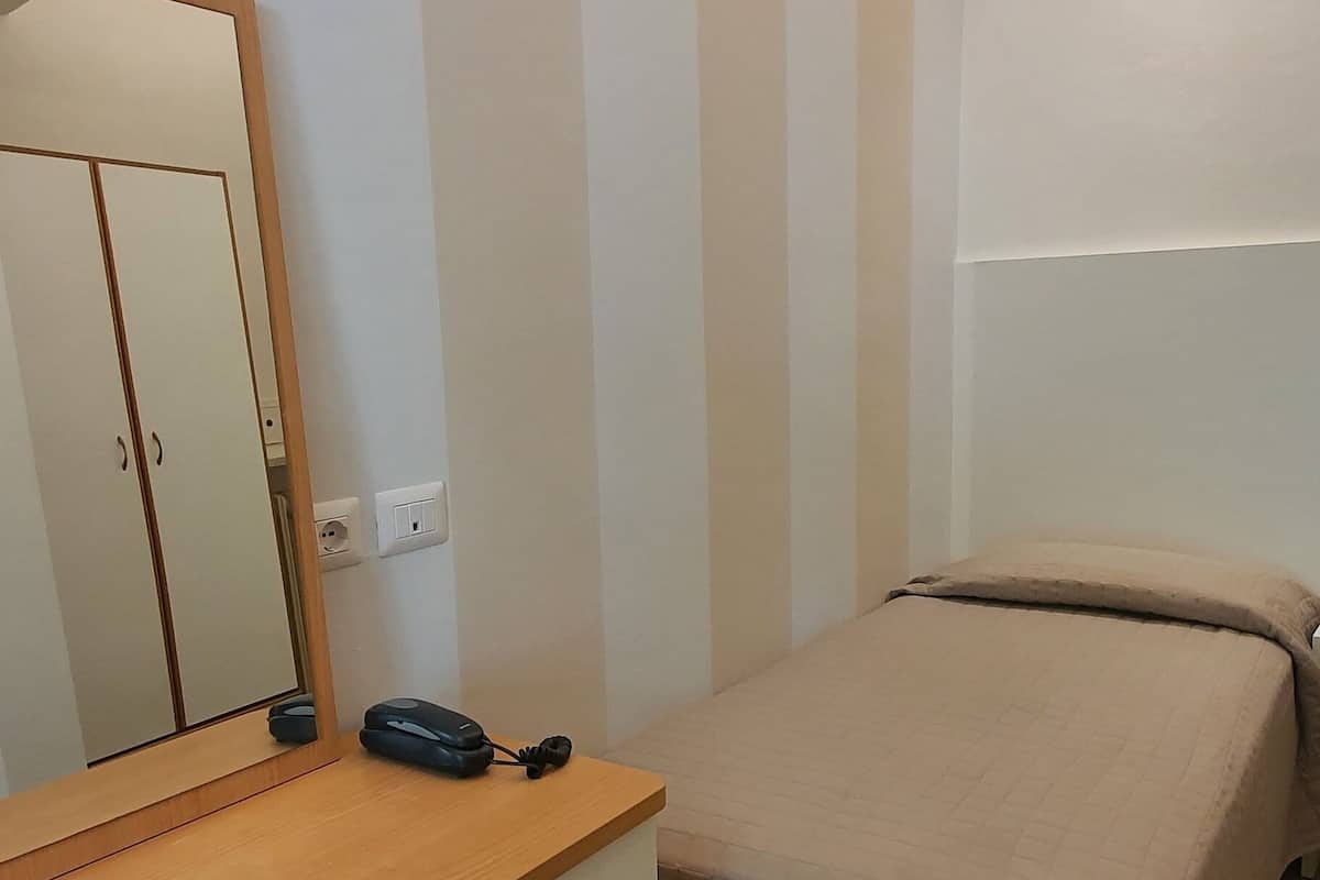 A bedroom with a wooden desk, a bed with a beige bedspread, a mirror, and a ceiling light.