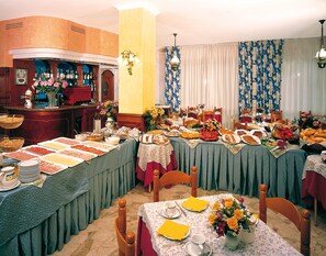 A dining area with multiple tables set for a meal, featuring a variety of dishes and a bar area in the background. - Hotel Napoleon (Jesolo)