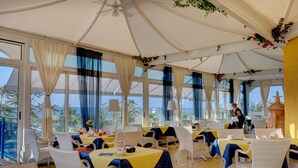 A restaurant with tables covered in yellow and blue tablecloths, white wicker chairs, and large windows offering a view of the sea. - Hotel Napoleon (Jesolo)