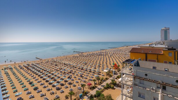 A beach with rows of sunbeds and umbrellas, a clear sky, and a building with a red roof in the background. - Hotel Napoleon (Jesolo)