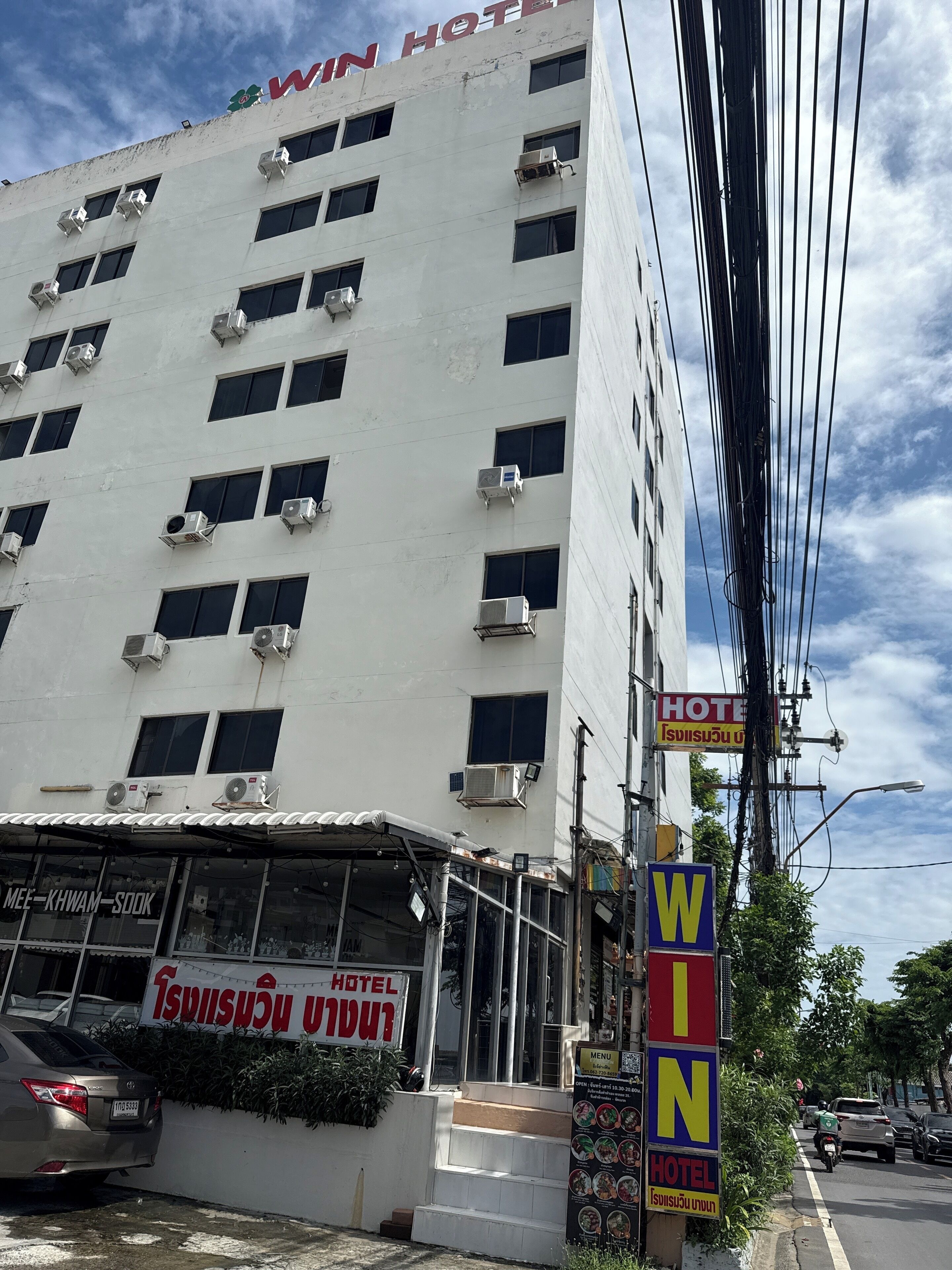 A hotel building with multiple air conditioning units, a signboard with Thai text, and a street view with parked cars and a motorcycle.
