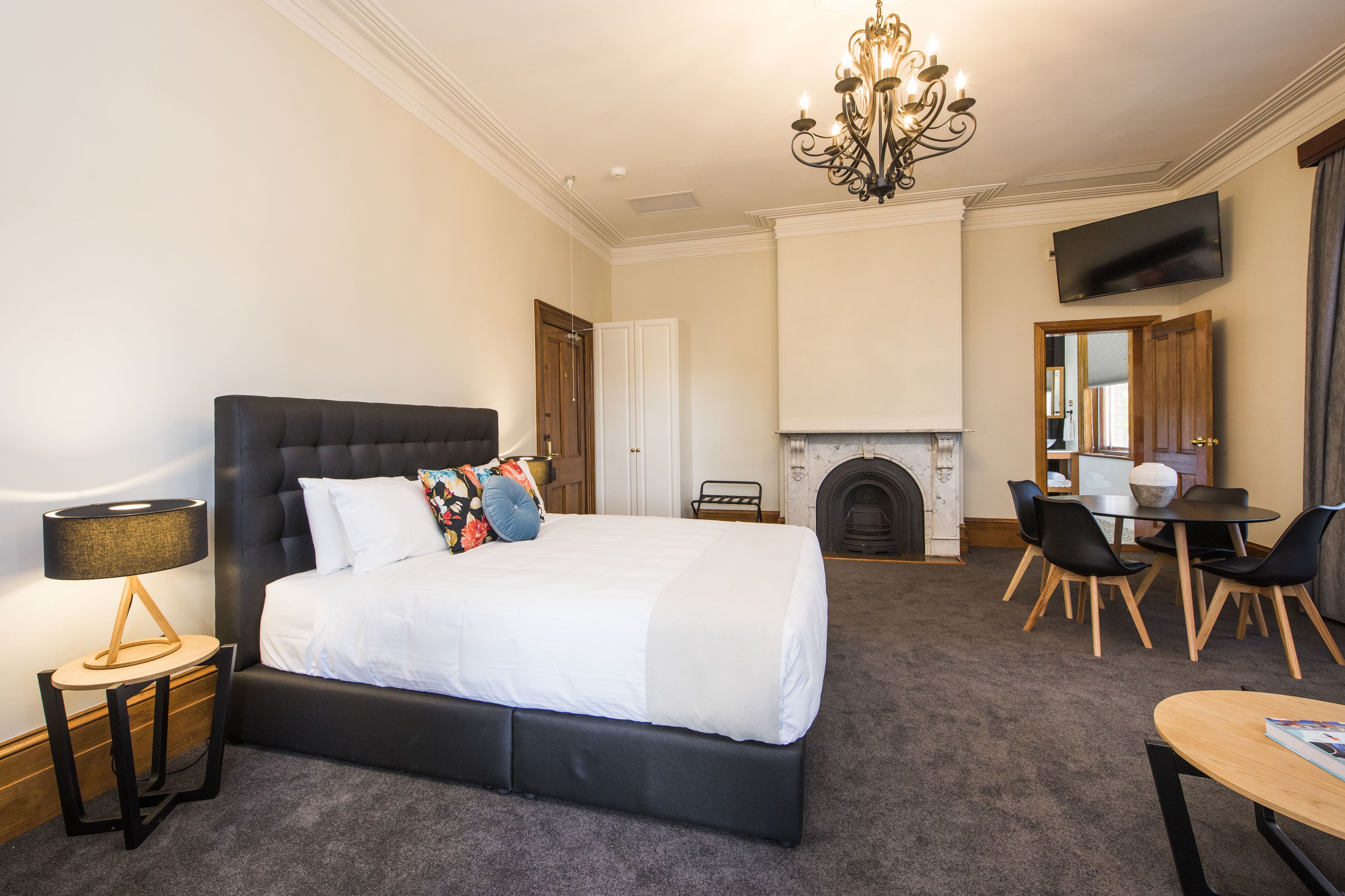 A bedroom with a large bed, a bedside table, a lamp, a fireplace, a TV, and a dining area with chairs and a table.