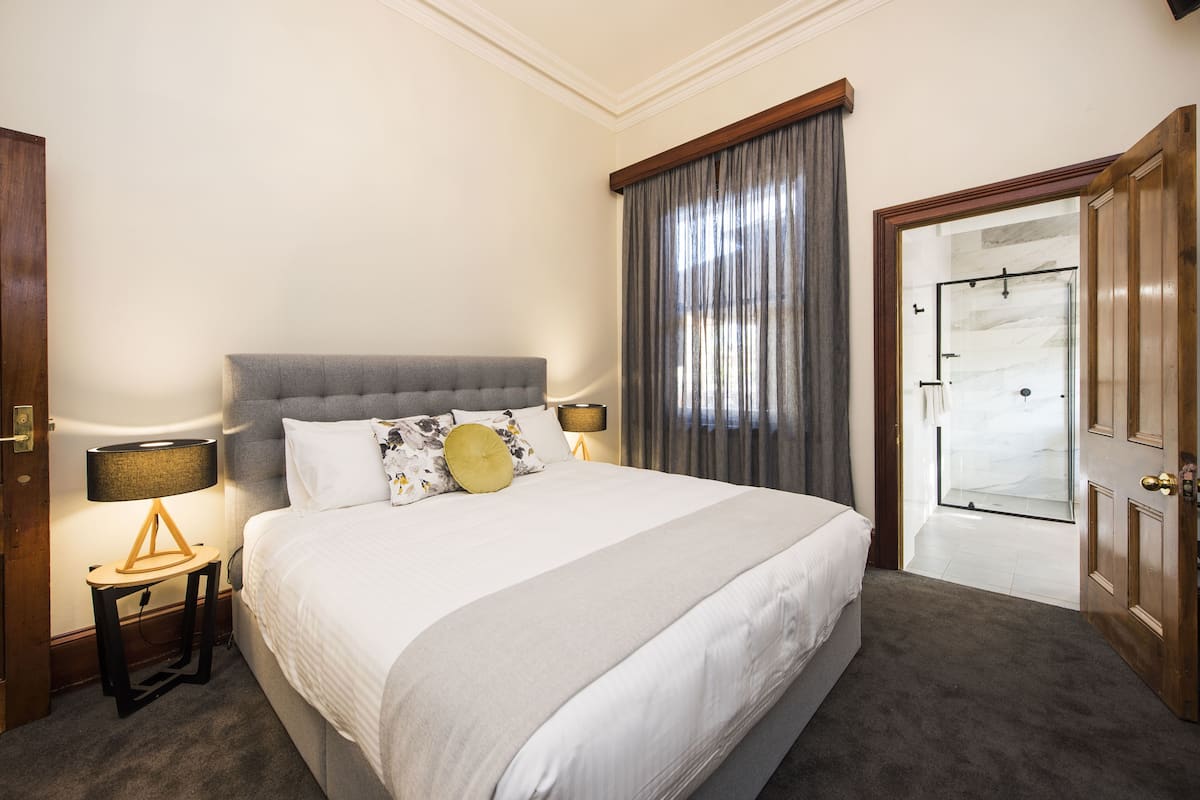 A neatly arranged bedroom with a large bed, two bedside tables with lamps, a window with curtains, and a door leading to another room.