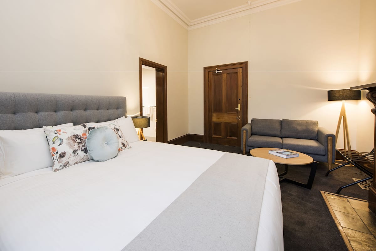A hotel room with a large bed, a grey sofa, a small round table, and a floor lamp.