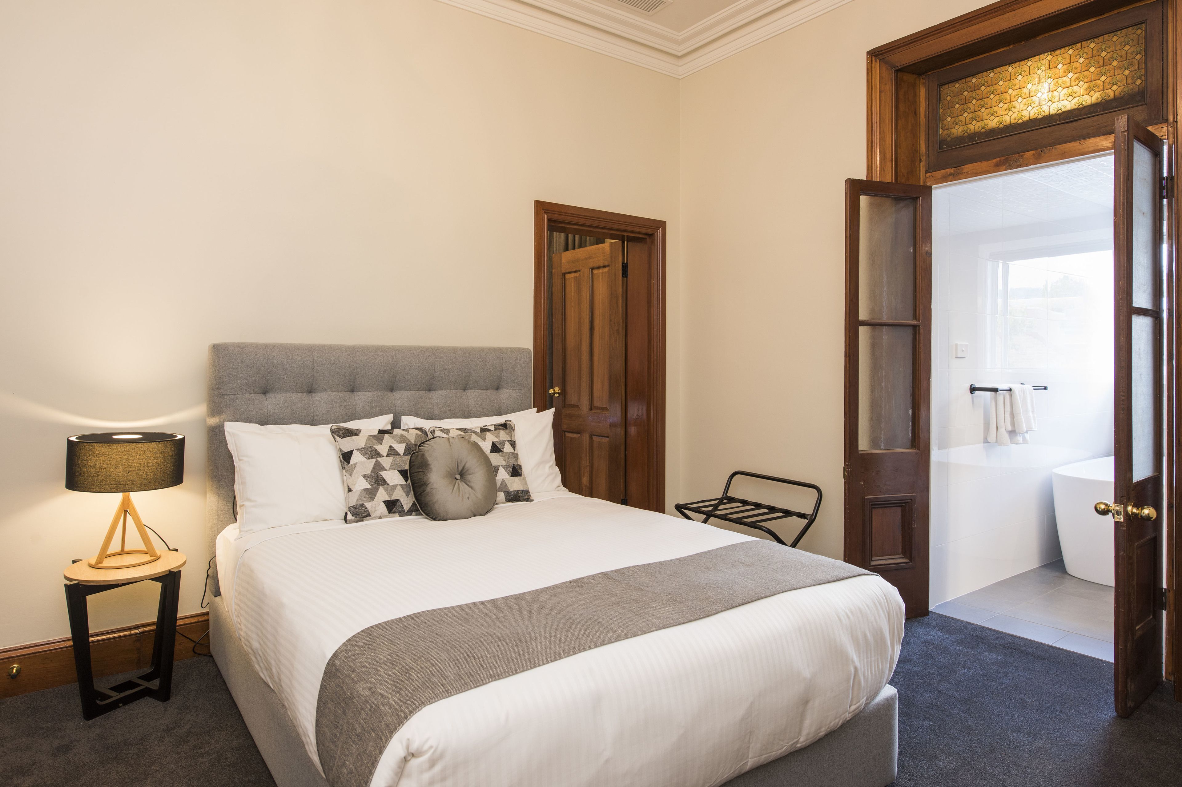 A neatly made bed with a grey headboard, a bedside lamp, and a small table with a lamp. A wooden door leads to a bathroom with a bathtub.