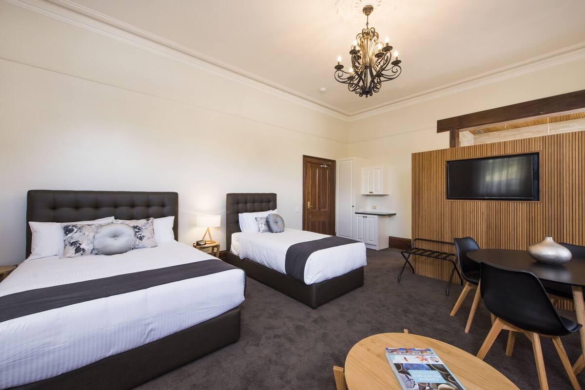 A hotel room with two beds, a desk, a chair, a TV, and a chandelier.