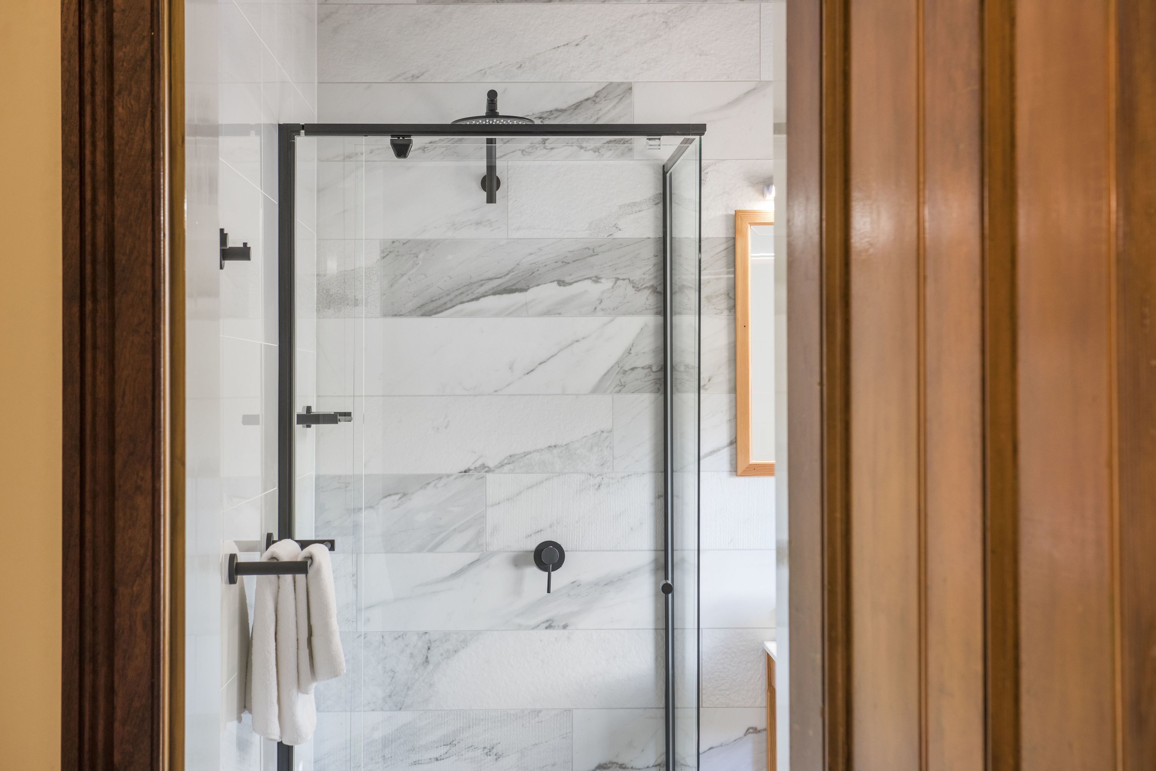 A glass-enclosed shower with a marble wall, a towel rack, and a wooden door frame.