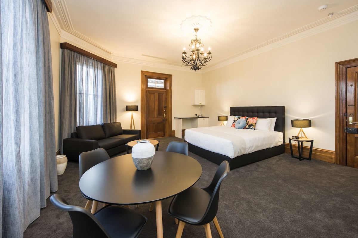 A hotel room with a bed, a sofa, a dining table with chairs, a chandelier, and a door.