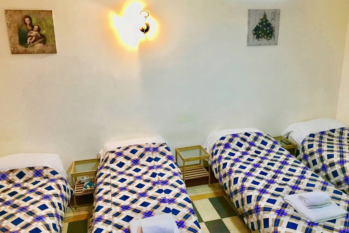 A room with four beds, each with a checkered bedspread, a nightstand, a wall-mounted light, and a framed picture on the wall.