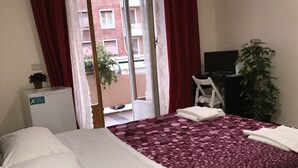 Une chambre d’hôtel avec un lit, un bureau, une chaise, un petit réfrigérateur et une plante en pot.