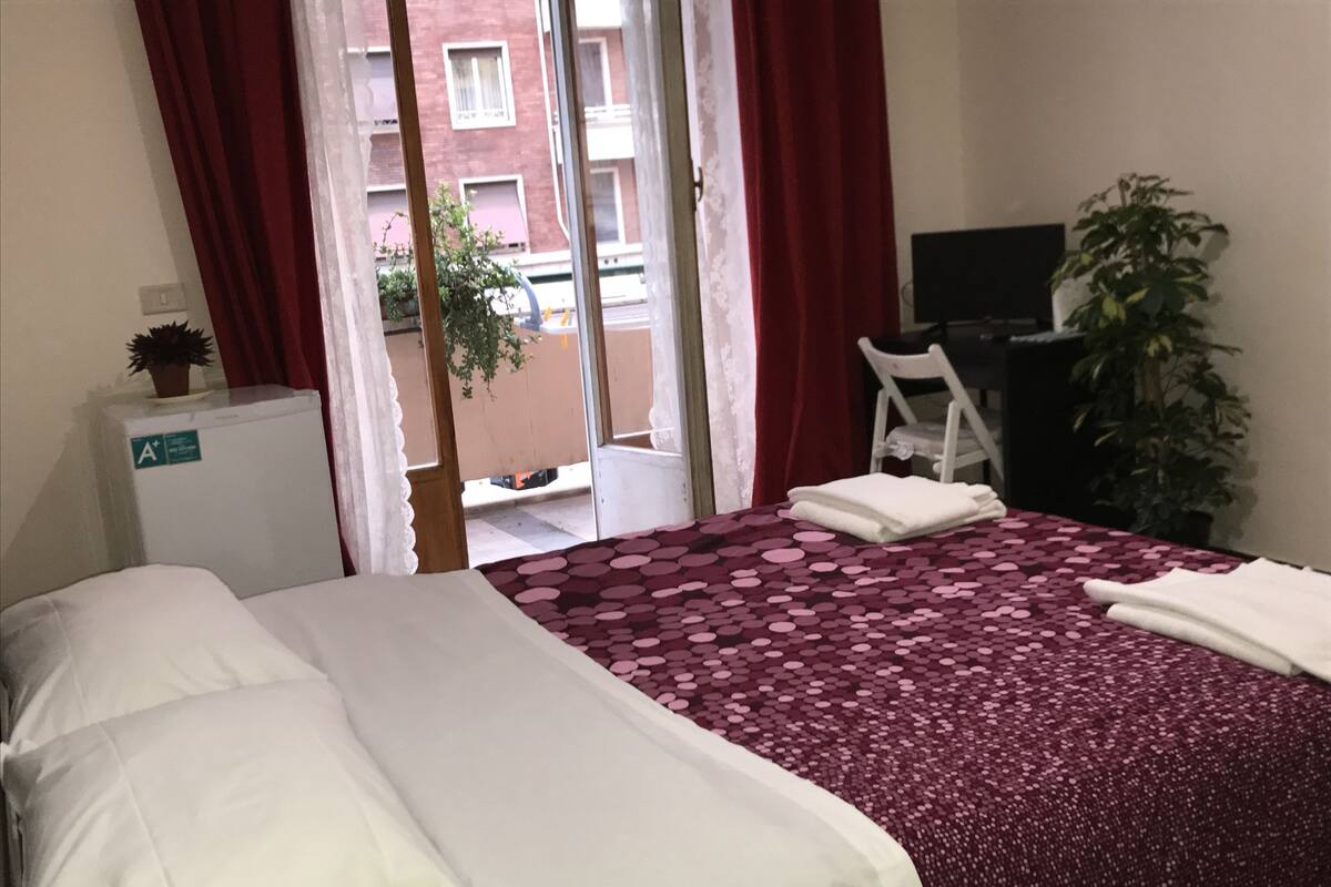 A hotel room with a bed, a desk, a chair, a small refrigerator, and a potted plant.
