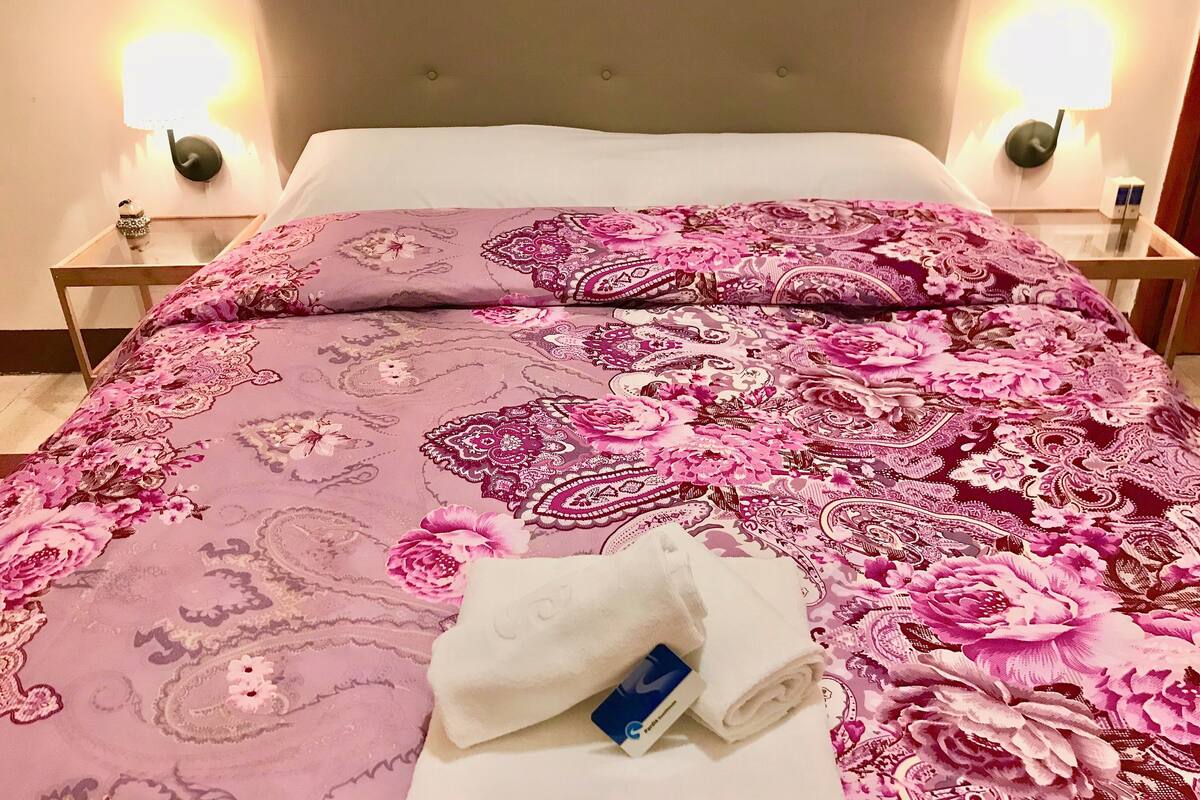 A bed with a floral-patterned bedspread, two bedside lamps, and a folded white towel with a blue tag on a wooden nightstand.