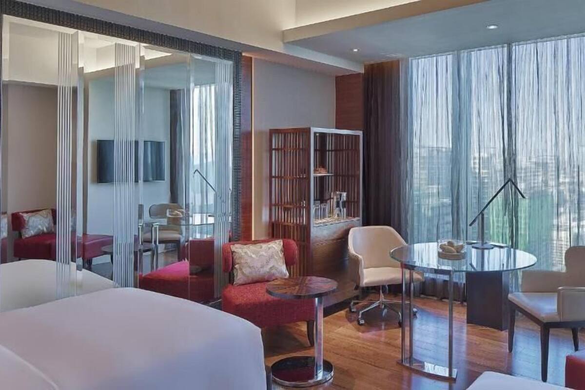 A modern hotel room with a glass partition, a round dining table, a bookshelf, and a large window with curtains.
