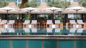 A pool area with lounge chairs and thatched umbrellas, surrounded by greenery and modern buildings.