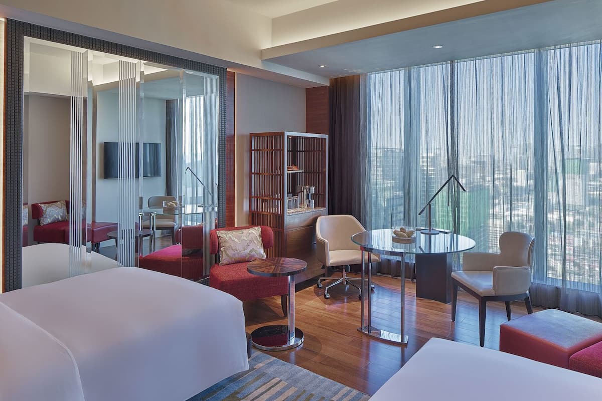A hotel room with a large bed, a desk, a chair, a sofa, a bookshelf, and a large window with curtains.