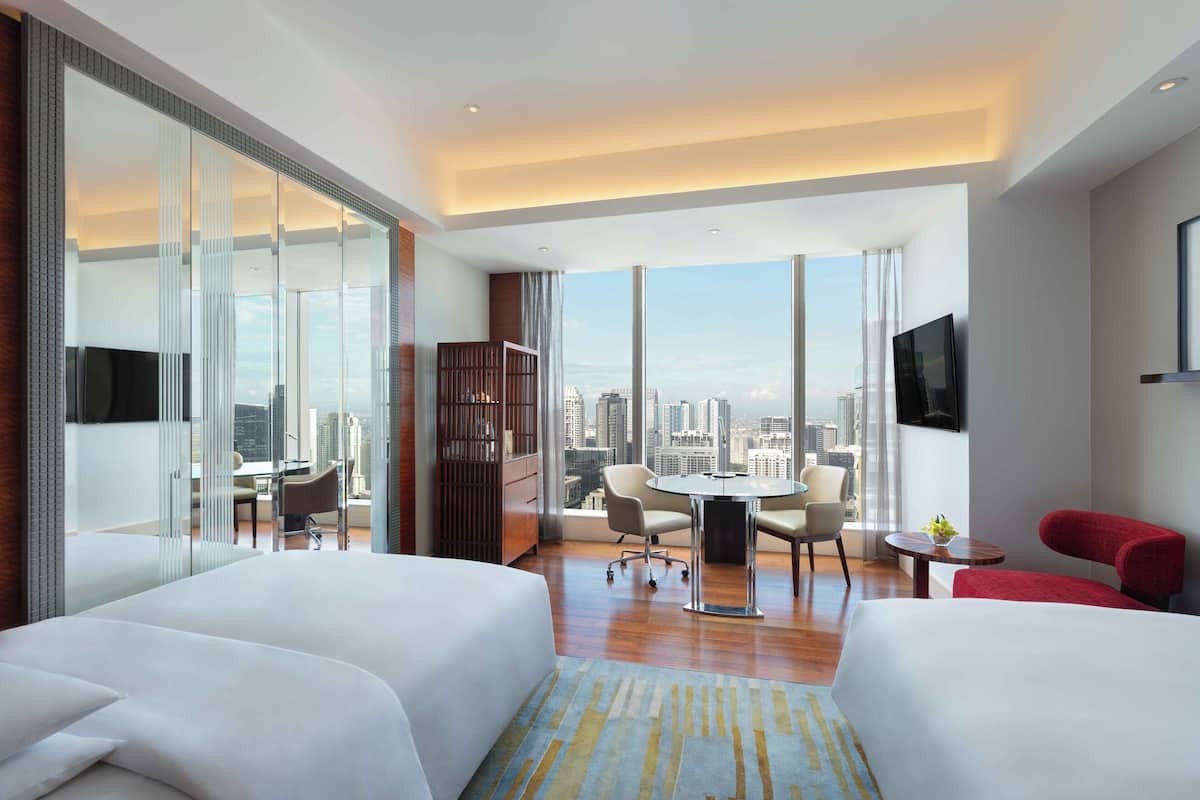 A modern hotel room with two beds, a dining area, a TV, and a city view.