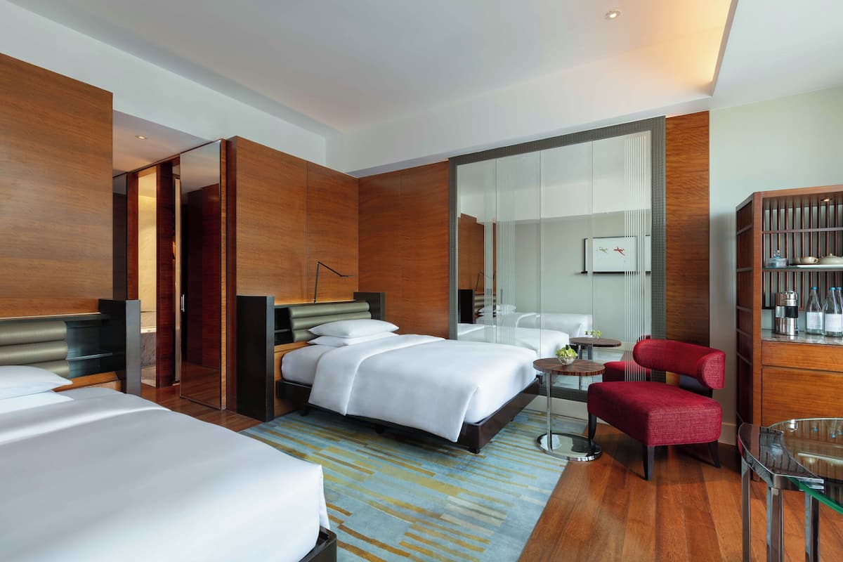 A modern hotel room with a large bed, a sitting area with a red chair, a small table, and a glass coffee table.
