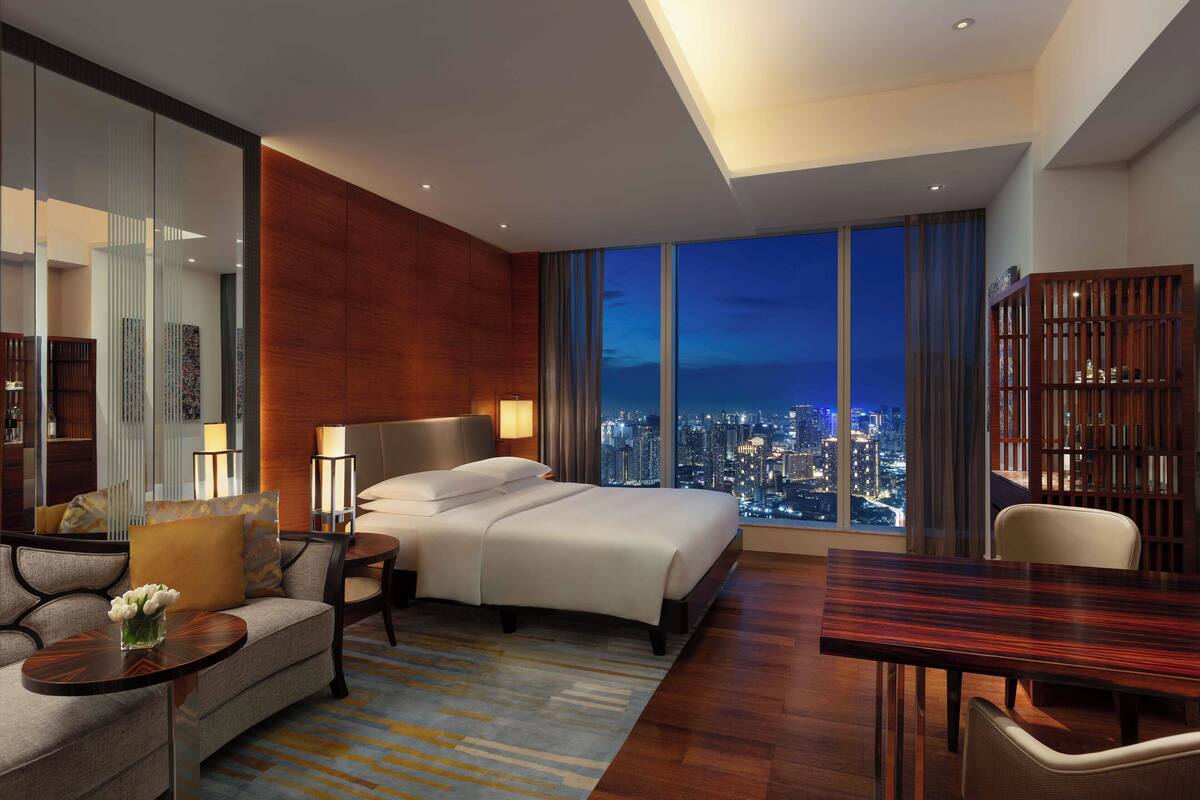 A modern hotel room with a large bed, a sofa, a dining table, and a city view.