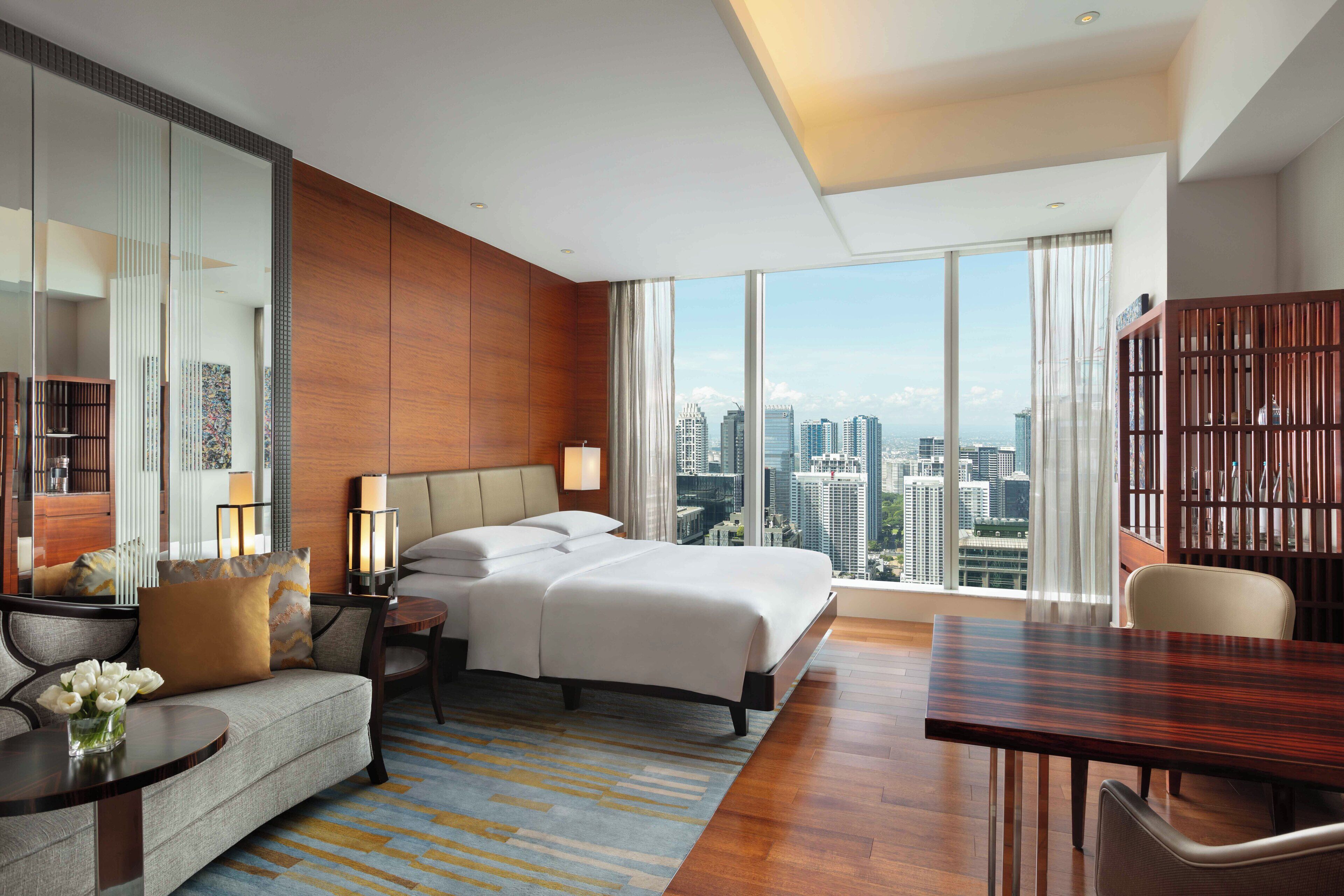 Quarto de hotel moderno com cama grande, sofá, mesa de jantar e vista para a cidade.