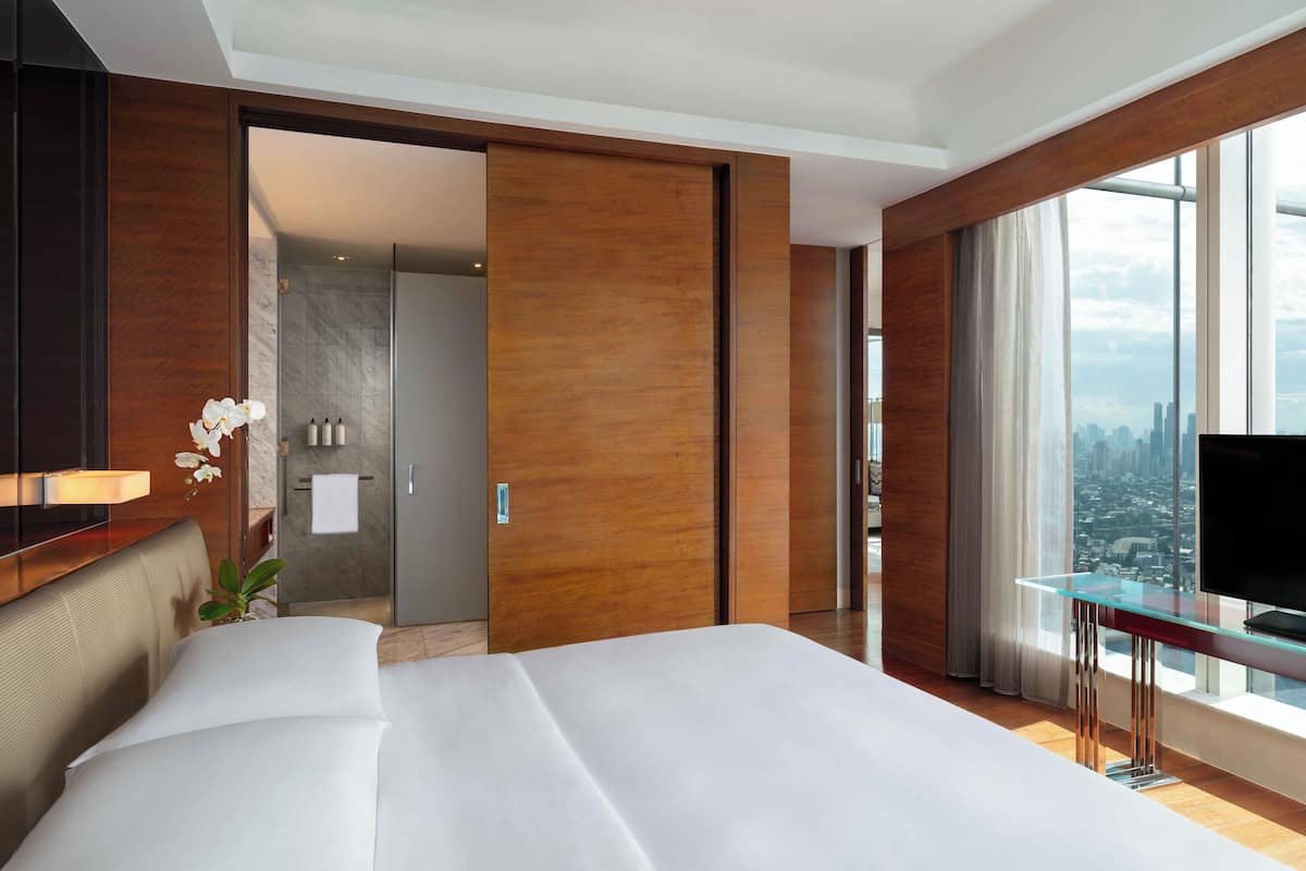 A modern hotel room with a large bed, a glass table, a TV, and a city view.