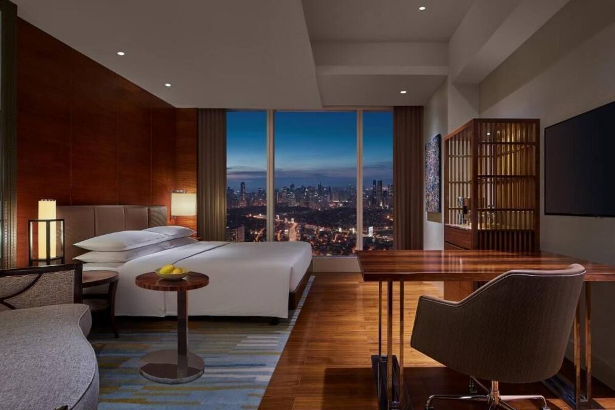 A modern hotel room with a large bed, a desk, a chair, a bookshelf, and a city view.