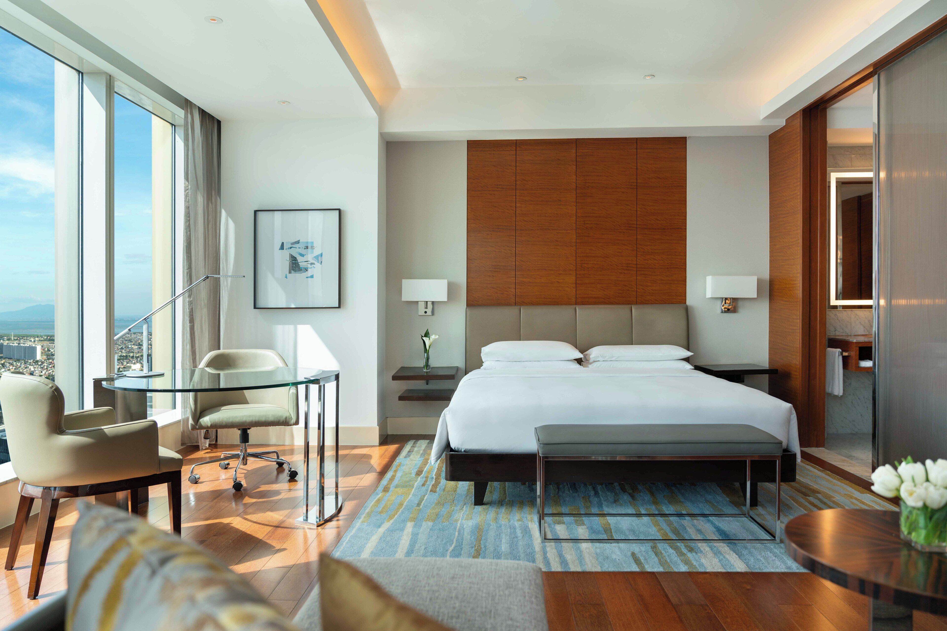 A modern hotel room with a large bed, a glass table, a chair, a sofa, and a view of the city.