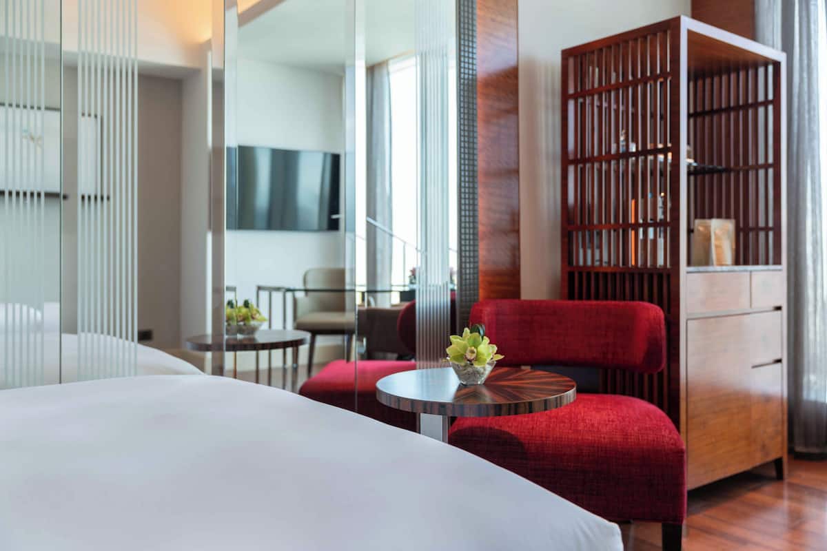 A modern hotel room with a large bed, a red armchair, a small round table with a vase of flowers, a wooden cabinet, and a TV mounted on the wall.