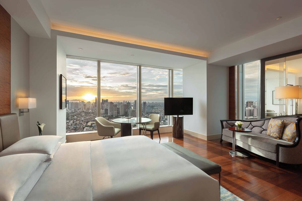 A hotel room with a large bed, a sofa, a TV, a desk, and a view of a cityscape.