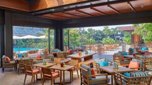 Un espacio de comedor al aire libre con mesas y sillas de madera, ventanas amplias y vistas a una piscina y follaje.