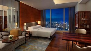 Habitación de hotel moderna con una cama grande, un sofá, una mesa de comedor y vistas a la ciudad al atardecer.
