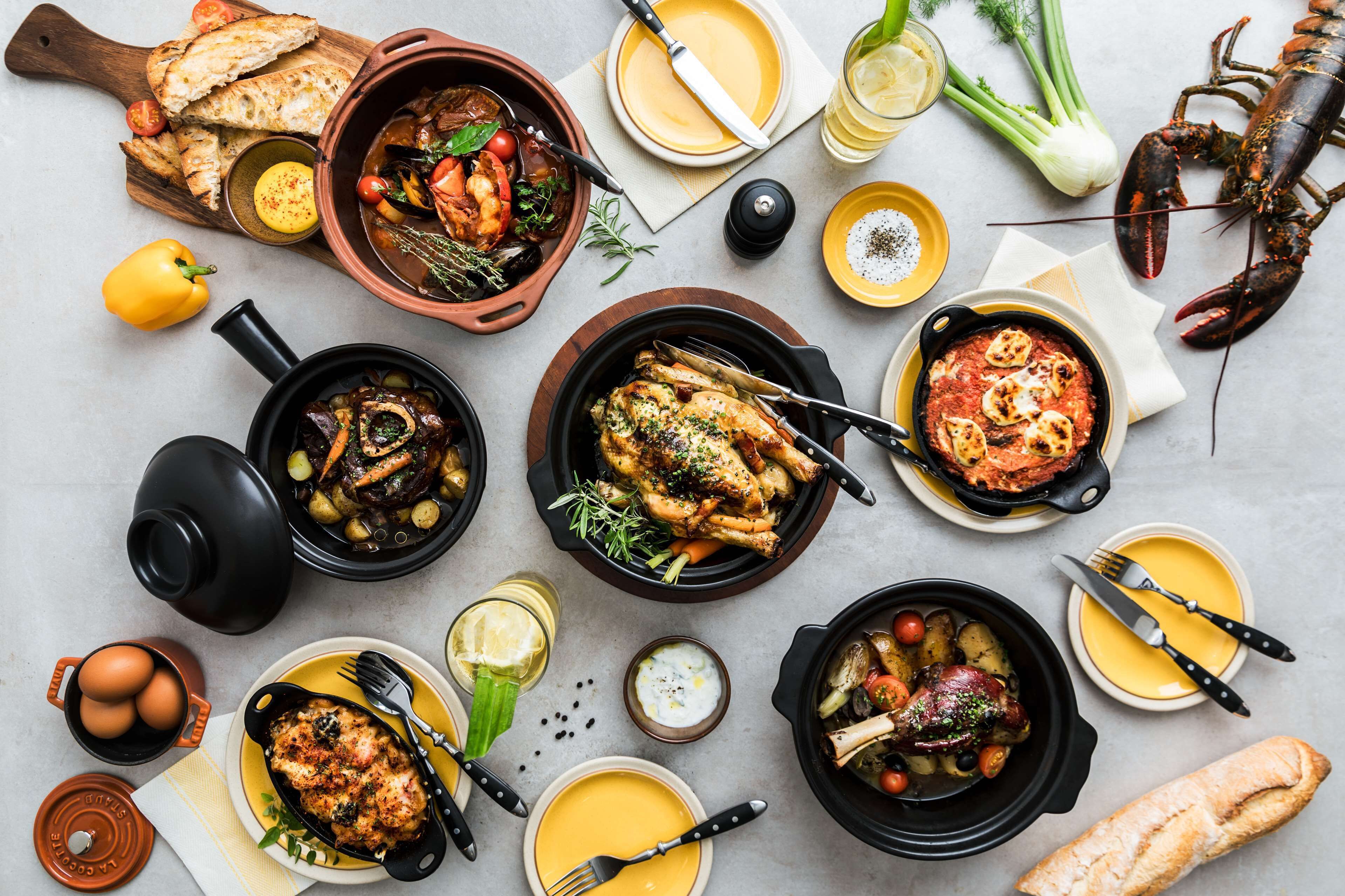 Variedad de platos que incluyen pollo asado, verduras y mariscos, dispuestos sobre una mesa con platos, cubiertos y aderezos.