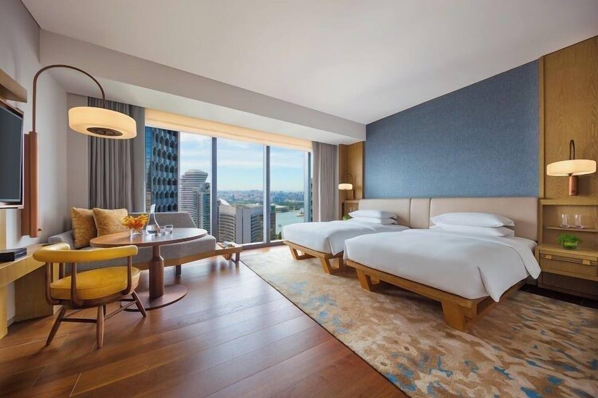 A hotel room with a large window, a sofa, a dining table, two beds, and a small side table.