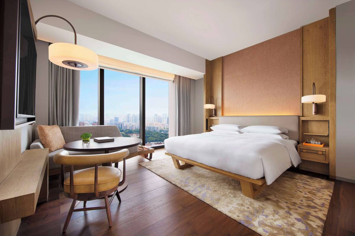 A modern hotel room with a large bed, a desk with a chair, a round table, and a city view.