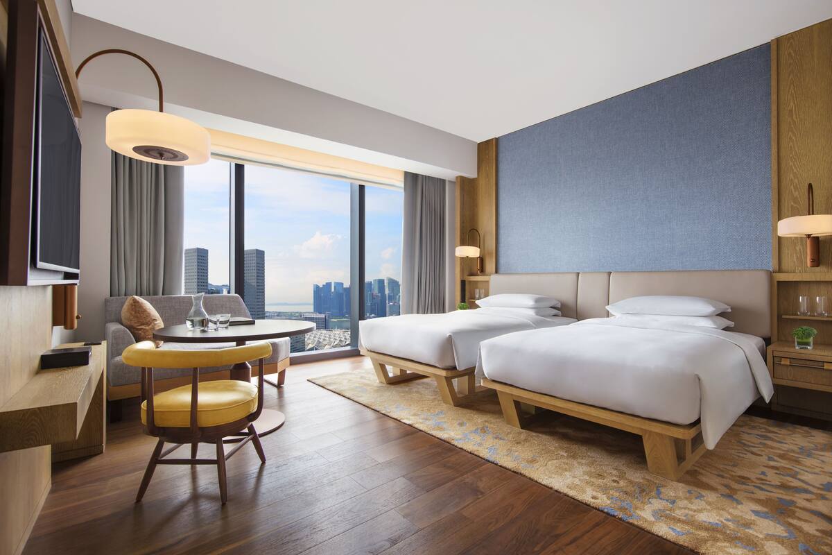 A hotel room with two beds, a desk, a chair, a large window with a city view, and a modern lamp.