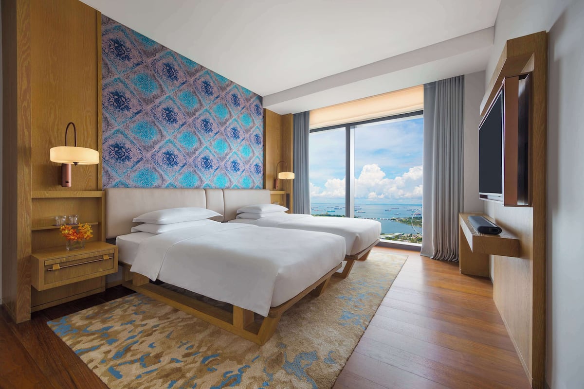 A hotel room with a large bed, a TV, a desk with a lamp, a view of the sea, and a patterned wall.
