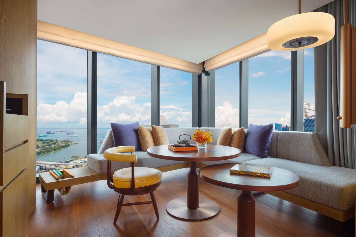 A modern hotel room with a large window offering a city view, a sofa, two round tables, and a small wooden side table.