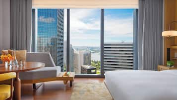 A hotel room with a large bed, a sofa, a dining table with a vase of flowers, and a view of city buildings and a river.