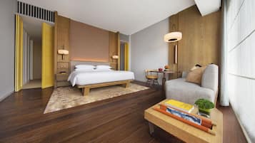 A modern hotel room with a wooden floor, a large bed, a sofa, a small table with books and a plant, and a desk with a lamp.