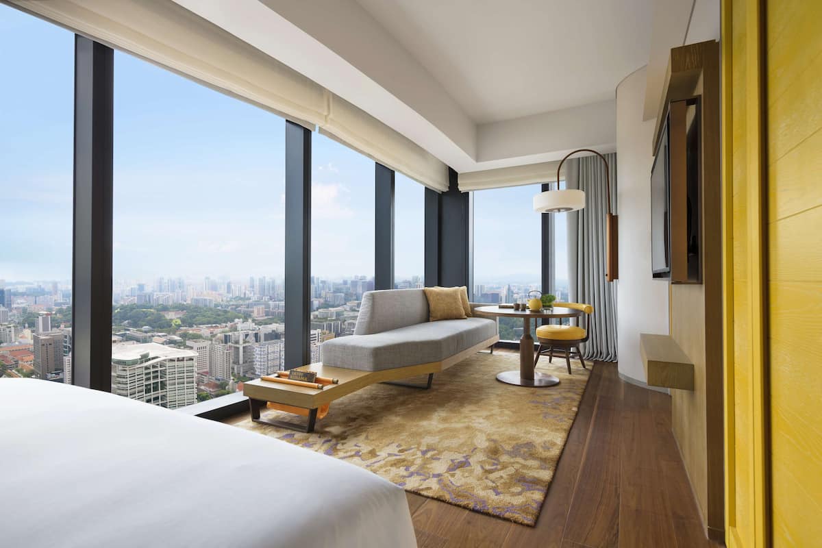 A modern hotel room with a large window offering a city view, a sofa, a small table with a lamp, and a bed.