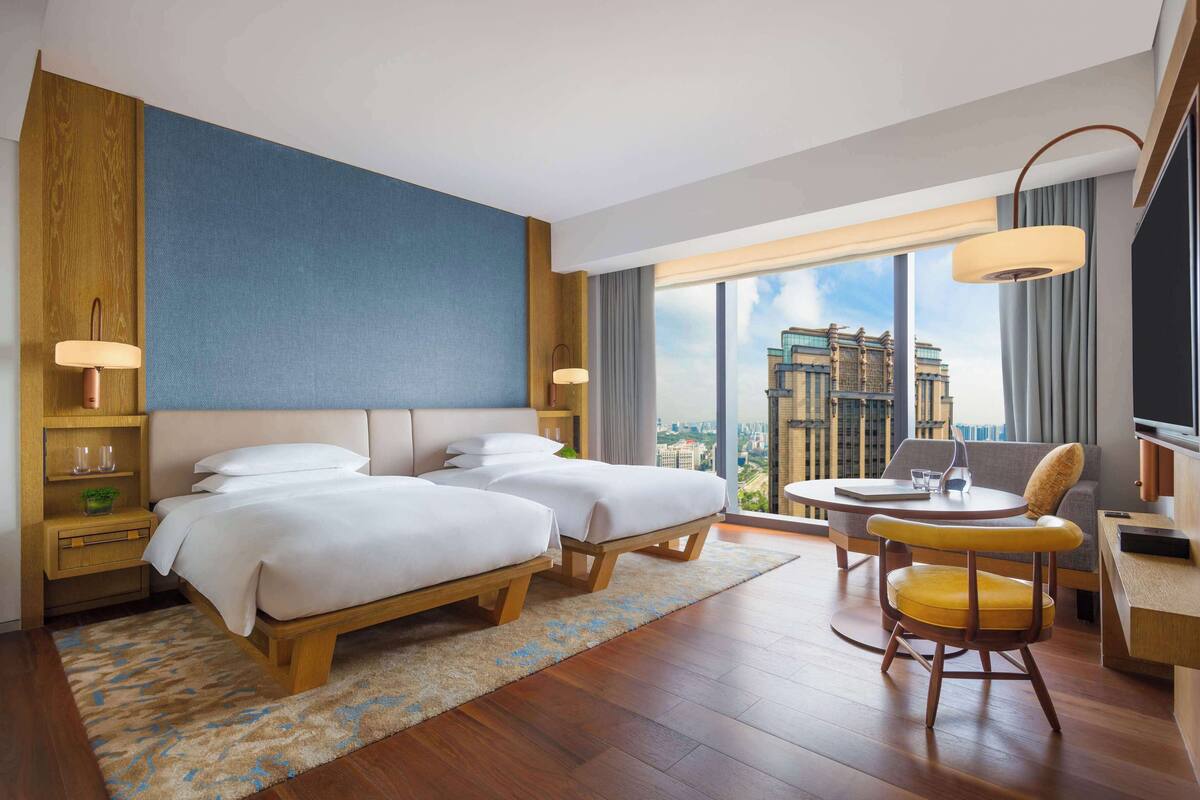 A modern hotel room with two beds, a desk, a chair, and a large window offering a city view.