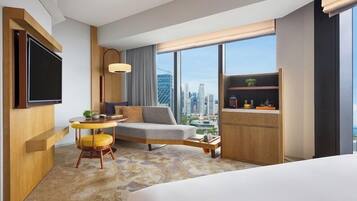 A modern hotel room with a large bed, a seating area with a sofa and chairs, a small table, a TV, and a view of the cityscape through large windows.