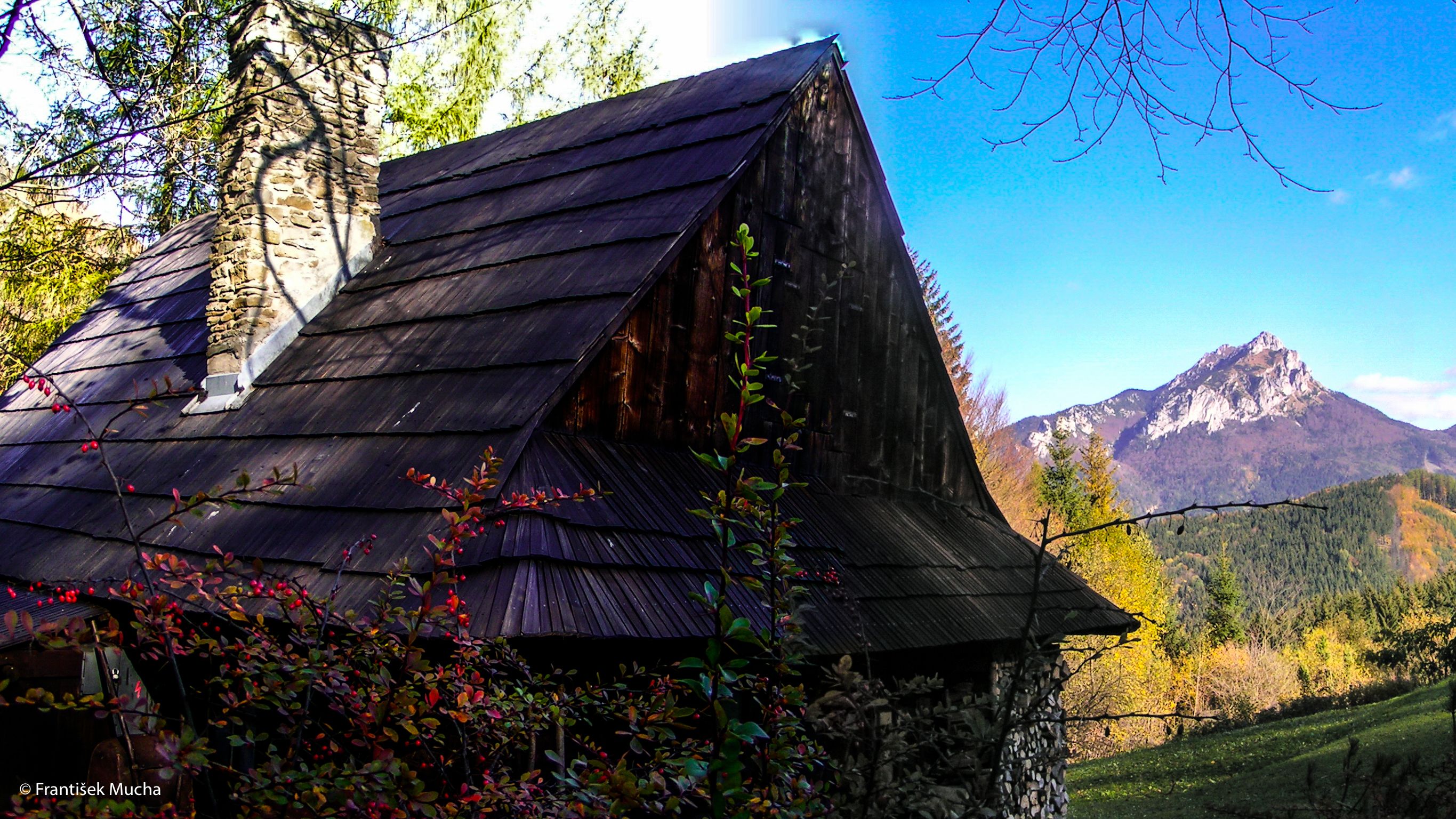 A rustic stone cabin with a steeply pitched roof, surrounded by autumn foliage and a mountain backdrop.