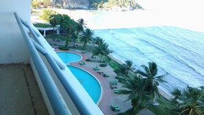 A pool with a curved edge, surrounded by palm trees and lounge chairs, overlooking a beach and ocean.