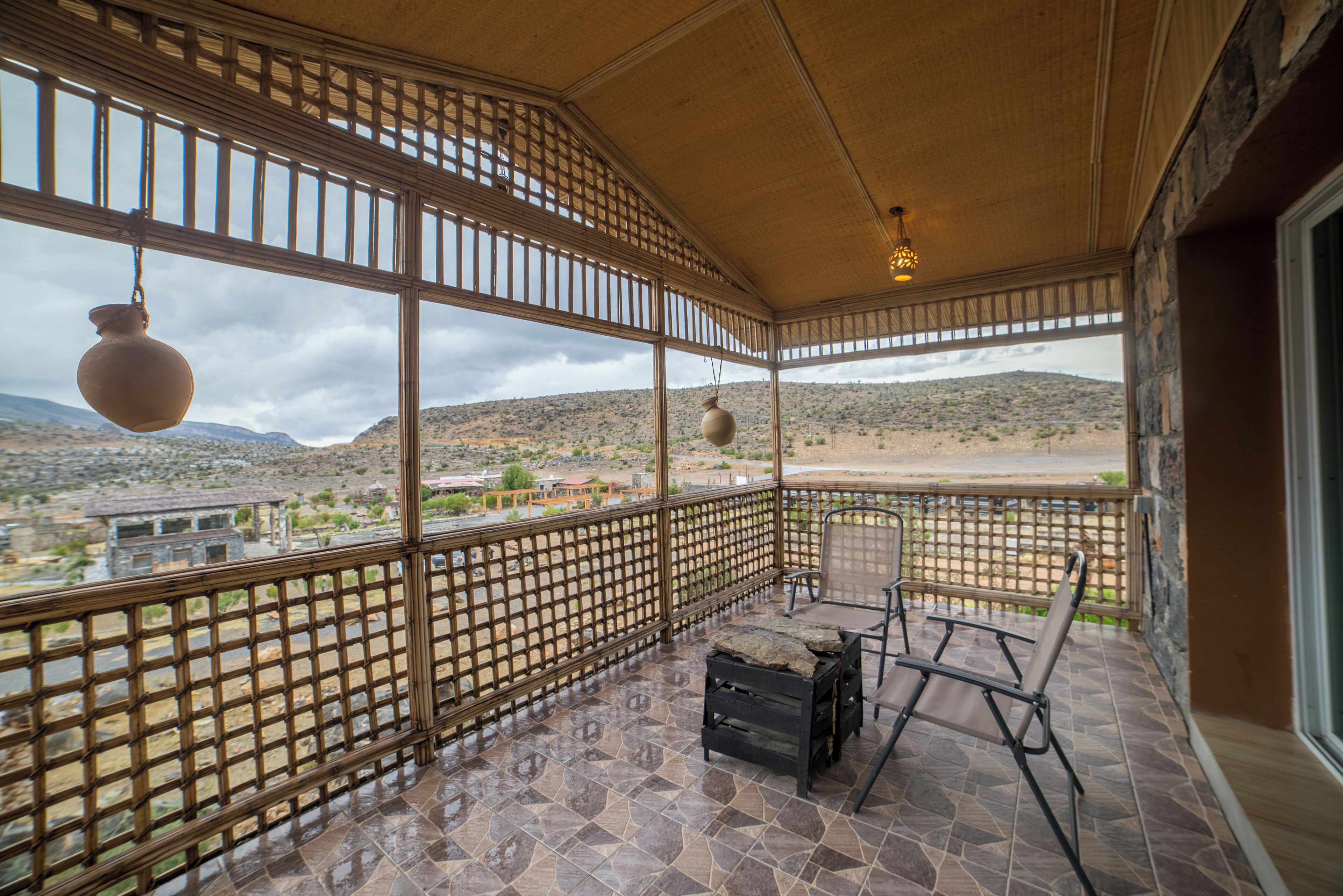 A covered patio with a view of a desert landscape, featuring a wooden lattice partition, a table with chairs, and hanging pottery.
